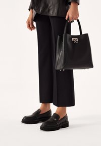 Schwarze Ledertasche mit strukturiertem Design, silberner Hardware und Logo-Akzent; kombiniert mit schwarzen Loafers und weitem Hosen.