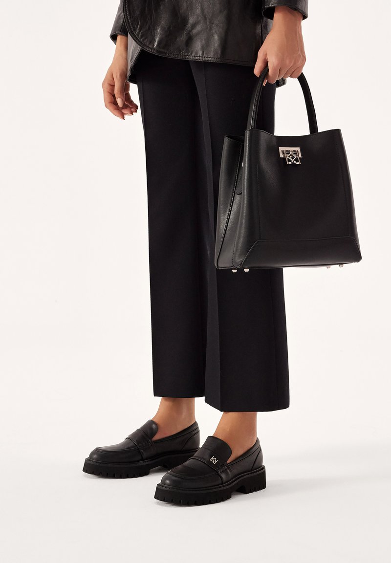 Schwarze Ledertasche mit strukturiertem Design, silberner Hardware und Logo-Akzent; kombiniert mit schwarzen Loafers und weitem Hosen.