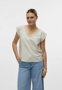Blusa beige chiaro con scollatura a V bordata di pizzo, maniche corte a cappuccio e vestibilità morbida. Indossata con jeans blu a vita alta.