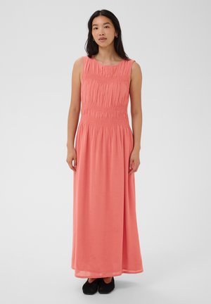 Donna in piedi che indossa un vestito lungo color corallo rosa senza maniche con corpetto arricciato e scarpe basse nere su uno sfondo chiaro e uniforme.
