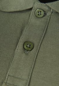 Oliwkowa koszulka polo o teksturowanej tkaninie, z zapięciem na guziki i haftowanym kołnierzykiem. Zbliżenie na guziki i detale listwy.