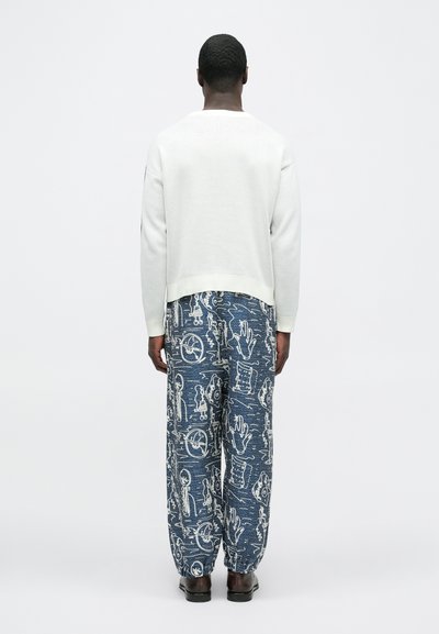 Λευκό φούτερ με υφή, συνδυασμένο με ανοιχτό μπλε πα ντελόνι με σχέδια που περιλαμβάνουν αφηρημένες λευκές εικόνες, φορεμένο με σκούρα παπούτσια.