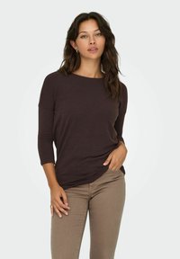 Maglietta a maniche lunghe marrone scuro con vestibilità rilassata, scollo rotondo e texture morbida, abbinata a jeans aderenti marrone chiaro.