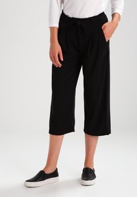 Pantalon court noir en tissu doux, avec taille à cordon de serrage et poches avant. Associé à des chaussures noires faciles à enfiler.