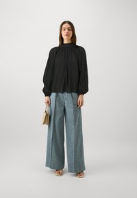 Blusa nera con scollo arricciato e maniche lunghe a palloncino, abbinata a pantaloni larghi blu chiaro e una piccola borsa beige.