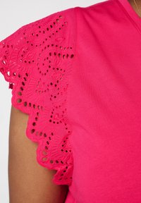 Roze top met korte mouwen, voorzien van een kanten rand aan de mouwen met ingewikkelde bloemmotieven en uitsnijdingen. Zachte, gladde textuur.