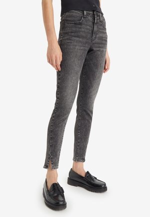 Personne portant un jean skinny noir délavé avec des fentes à l'avant aux chevilles et des mocassins noirs, debout devant un fond blanc.