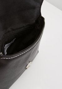 Vnitřek černé kožené tašky, se zipovou kapsou, hladkým povrchem a stříbrným kováním. Odhalené stehy zdůrazňují detail designu.