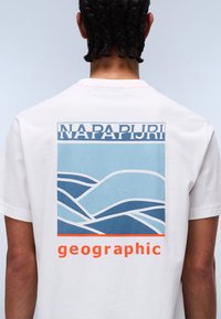 Bílé bavlněné tričko s modrou vlnkovou grafikou a oranžovým textem "geographic" a "NAPAPIJRI" nahoře. Krátké rukávy a kulatý výstřih.
