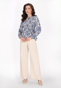 Leichte Bluse mit blauem Blumenmuster auf cremefarbenem Hintergrund, kombiniert mit beigen weiten Hosen und nude-farbenen High Heels.