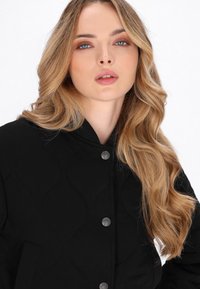 Blonde Frau mit welligem Haar trägt eine schwarze gesteppte Jacke mit Knopfverschluss und posiert vor einem schlichten weißen Hintergrund.
