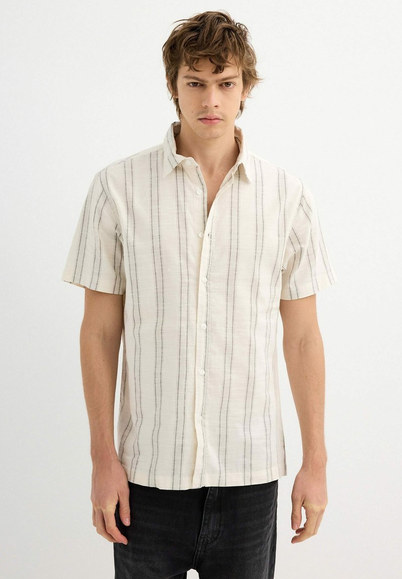 Camisa de manga corta en tejido crema claro con rayas verticales negras, con un cierre de botones en la parte delantera y un ajuste relajado.