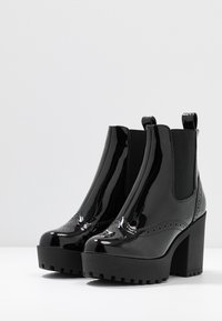 Botines de charol negro con tacón alto de bloque, paneles laterales elásticos y suela de goma texturizada. Detalles clásicos de brogue en la parte frontal.
