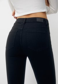 Persona con jeans ajustados negros de cintura alta y una camiseta blanca de manga larga recortada, vista desde atrás sobre un fondo liso.