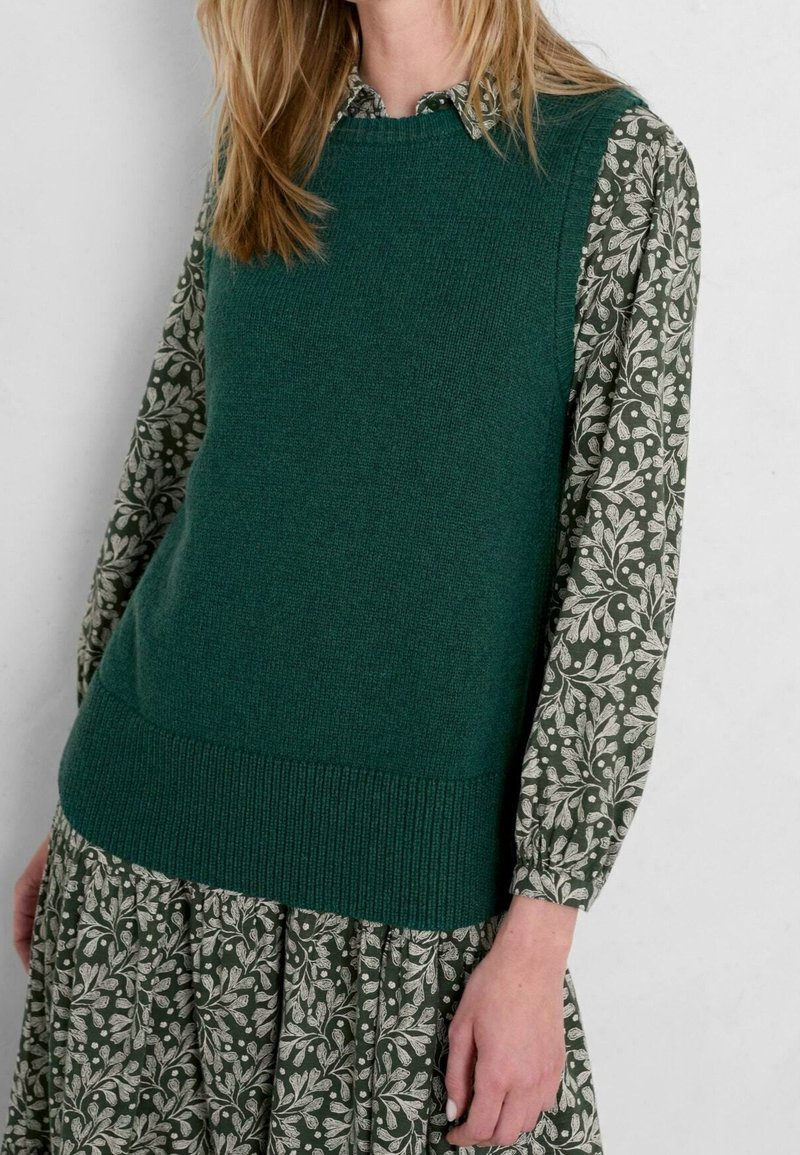 Grüner Strickpullover mit rundem Ausschnitt und strukturierter Stoffstruktur, kombiniert mit einem Kleid mit Blumenmuster in Grüntönen und Cremefarben.