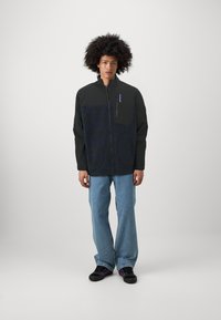 Giacca nera con zip caratterizzata da un motivo grafico blu tessurizzato sul petto, con una tasca con zip sul lato sinistro. Indossata con jeans azzurri chiari.