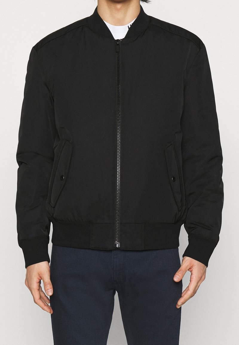 Giacca bomber nera con zip frontale, tasche laterali, polsini e orlo a costine. Realizzata in tessuto liscio con un design semplice e classico.