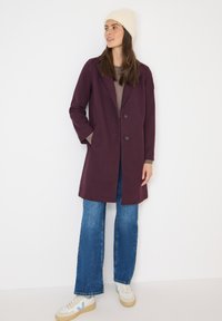Cappotto in bordeaux con rever a punta e due bottoni, indossato sopra un maglione a maglia beige, abbinato a jeans svasati e sneakers beige.