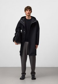 Svart vattentät jacka med justerbar huva, framknäppning och stora fickor. Modellen håller en svart handväska och är klädd i grå byxor och skor.