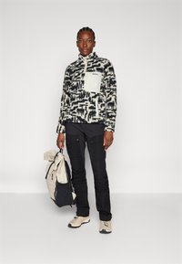 Giacca in fleece con motivo astratto bianco e nero, chiusura a zip e tasca sul petto; abbinata a pantaloni neri e uno zaino beige.