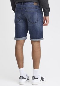 Tamne traperice u tamnoplavoj boji s okrenutim manžetama, prikazuju stražnje džepove. Uparene s bijelim Adidas tenisicama i čarapama. Minimalistički dizajn.