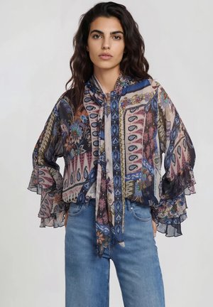 Frau mit langen dunklen Haaren trägt eine bunte, transparente Bluse mit Blumen- und geometrischen Mustern, kombiniert mit blauen, hoch taillierten Jeans.