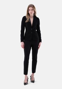 Schwarzes Damenanzug bestehend aus einem maßgeschneiderten Blazer und schmalen Hosen. Der Blazer hat zwei Knöpfe, ausgeprägte Revers und einen strukturierten Stoff.