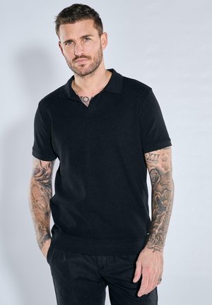 Schwarz Poloshirt mit kurzen Ärmeln, gerippter Textur, flachem Kragen und lockerer Passform, kombiniert mit dunklen Hosen.