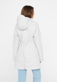 Kobieta stoi tyłem, ubrana w dopasowaną, jasnoszara kurtkę z kapturem i jasnoniebieskie dżinsy na białym tle.