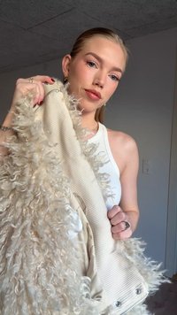 Beige jacket with textured, curly faux fur trim; ribbed fabric at the body; silver zipper; worn by a model in a casual setting.