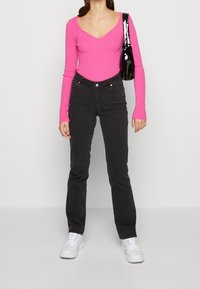 Růžové žebrované tričko s dlouhým rukávem a výstřihem do V, doplněné černými džíny s vysokým pasem a bílými teniskami. Modelka drží lesklou černou kabelku.