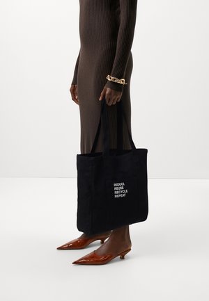 Personne portant une robe marron côtelée et un bracelet chaîne en or tient un sac cabas noir avec le texte blanc « Réduire Réutiliser Recycler Répéter ».