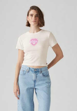 T-shirt corto a maniche corte color crema con un logo a forma di cuore rosa e il nome del marchio "Levi's." Abbinato a jeans a vita alta di colore blu chiaro.