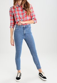 Camisa de cuadros rojos de botones con mangas remangadas, combinada con jeans ajustados de talle alto en azul y zapatillas negras con detalles en blanco.