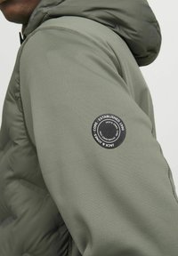 Oliven grøn pufferjakke med en glat tekstur, justerbar hætte og et cirkulært sort logo-patch på venstre skulder.