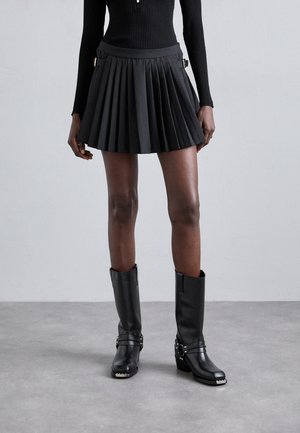 Plissé zwarte mini-rok gemaakt van gladde stof met zijzakken, gecombineerd met kniehoogte zwarte leren laarzen met metalen accenten.