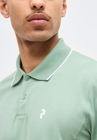 Άνδρας που φορά ανοιχτοπράσινο πόλο με λευκό περίγραμμα στο γιακά και μικρό λευκό λογότυπο στο στήθος, κοιτάζοντας ελαφρώς προς τα δεξιά σε απλό φόντο.