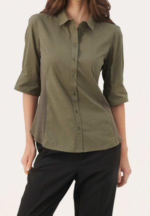 Frau trägt ein olivgrünes Hemd mit 3/4-Ärmeln und schwarze Hose, steht vor hellem, einfarbigem Hintergrund.