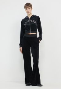 Donna che indossa un hoodie in velluto nero con la scritta "Angels" e pantaloni ampia coordinati, in piedi davanti a uno sfondo bianco uniforme.
