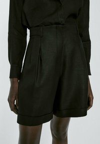 Shorts noirs taille haute en tissu texturé, avec des plis sur le devant et un ourlet replié, ornés de pièces métalliques à la taille.