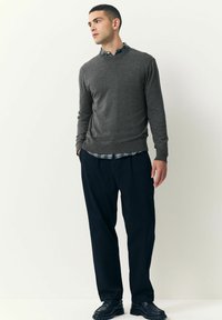 Joven con un suéter gris sobre una camisa de cuadros, con pantalones y zapatos negros, de pie con las manos en los bolsillos, mirando hacia la izquierda.