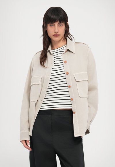 Μπεζ μπουφάν με εμπρός κλείσιμο με κουμπιά, δύο τσέπες στο στήθος και καφέ κουμπιά. Φορώντας πάνω από μια ριγέ ασπρόμαυρη μπλούζα και μαύρο παντελόνι.