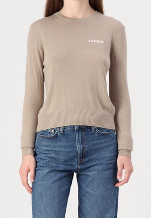 Person iført en beige langærmet sweater med "2NDDAY" logo på brystet og blå jeans, stående foran en ensfarvet baggrund.