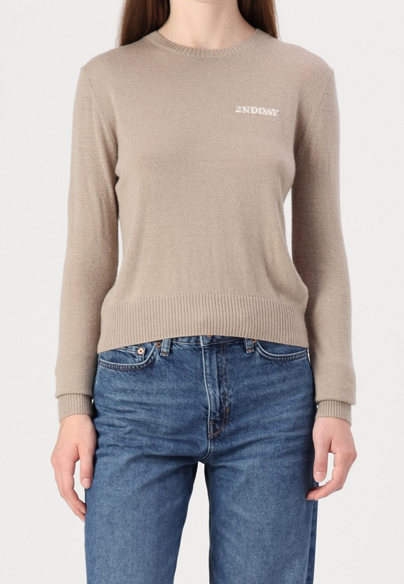 Personne portant un pull beige à manches longues avec le logo "2NDDAY" sur la poitrine et un jean bleu, debout devant un fond uni.