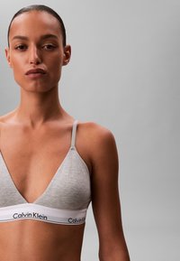 Jonge vrouw met donker haar in een staart, gekleed in een grijze Calvin Klein bralette tegen een effen grijze achtergrond.