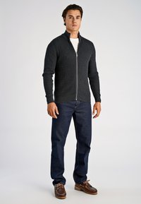 Dunkelgrauer Reißverschluss-Pullover mit strukturiertem Muster, getragen über einem weißen Hemd und kombiniert mit dunkelblauen Jeans und braunen Schuhen.