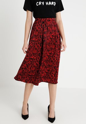 Kobieta ubrana w czerwoną i czarną spódnicę midi w wzory, czarne szpiczaste szpilki oraz czarny T-shirt z napisem "CRY HARD".