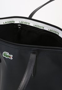 Svart tygväska med texturerat material, med ett vitt innerfoder med grön "LACOSTE"-logotyp och en liten grön krokodil-logga.