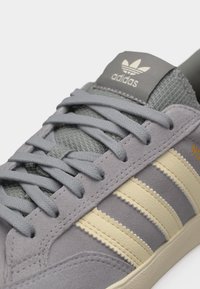 Κοντινή λήψη αθλητικού παπουτσιού Adidas σε γκρι χρώμα με γκρι κορδόνια, κρεμ τρεις ρίγες και λογότυπο Adidas στη γλώσσα.