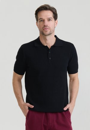 Mann mit kurzen braunen Haaren, der ein schwarzes kurzärmeliges Poloshirt und bordeauxrote Hose trägt, steht mit Händen in den Taschen vor einem schlichten Hintergrund.